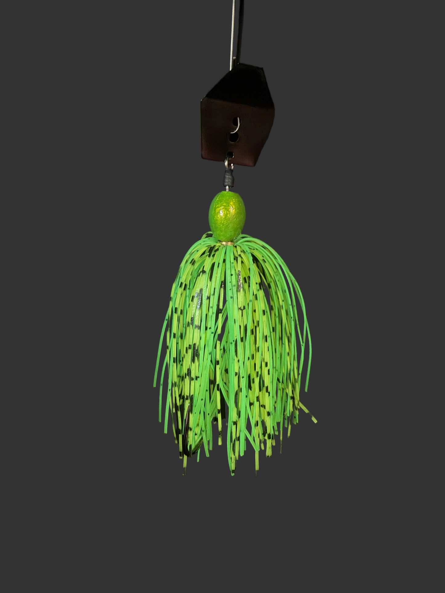 Green fishing lure with black speckles hanging on a hook against a dark background. 3/8 oz Chatterbait in Green and Lime Green with black speckles, wired skirt, and a #4/0 hook. Built in Canada to provoke aggressive bass and pike strikes.