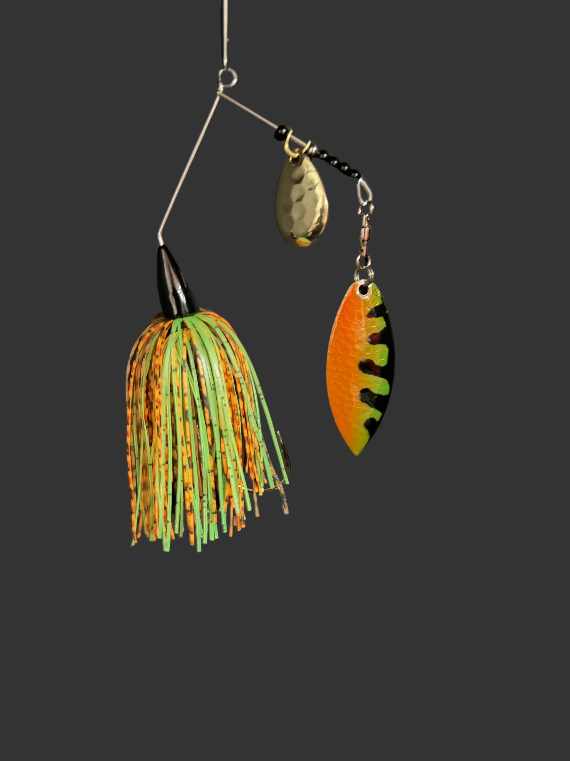 A fishing lure set hanging from a metal rig against a dark gray background. The set includes a large green and orange skirted jig, a small metallic teardrop-shaped lure, and an orange and black winged spinnerbait.