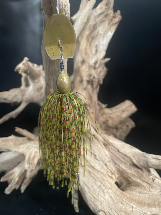 Fishing lure with a green and red pattern on a wooden surface. 3/8oz Chatterbait in Olive green with red shimmer & black speckles, wired skirt, and #4/0 hook. Mimics injured prey to trigger aggressive bass and pike strikes.