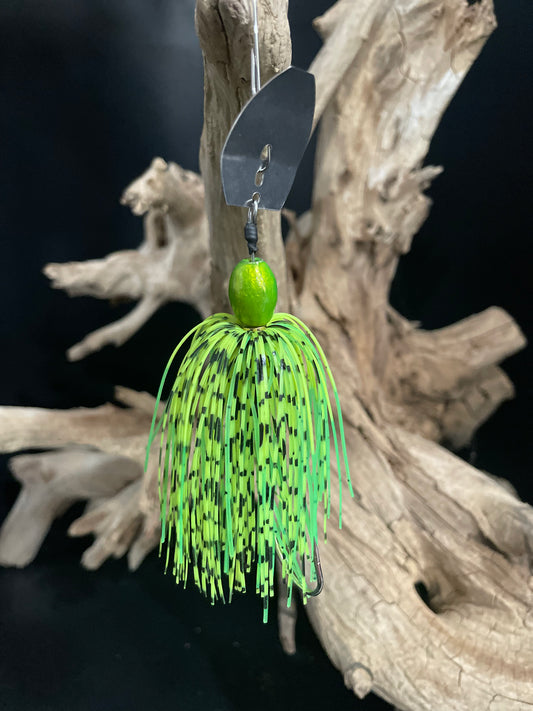 Green fishing lure with black speckles hanging on a hook against a dark background. 3/8 oz Chatterbait in Green and Lime Green with black speckles, wired skirt, and a #4/0 hook. Built in Canada to provoke aggressive bass and pike strikes.
