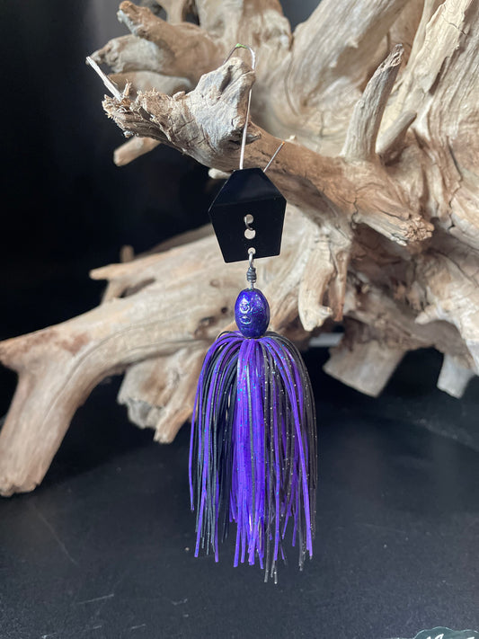 Purple and black Chatterbait fishing lure with a house-shaped top on a dark surface with driftwood. Lure-cifer